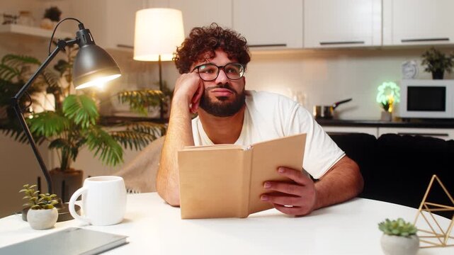 Arabian man at home reads dull book eyes heavy leans on hand slow page turn bored. Egyptian guy sitting at table keeps reading to finish chapter tries focus blinks often breath calm quiet night still
