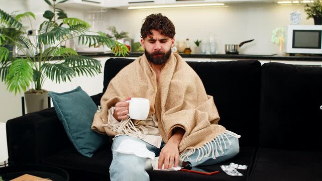 Indian man at home wrapped in blanket looks at smartphone reading health tips from doctor feels weak. Arabian guy on sofa holds tissue by nose cup of tea and pills near hand checks mobile phone sadly.