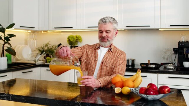Middle-aged man pours fresh orange juice then drinks with pleasure in home kitchen for healthy nutrition. Mature guy smiling after morning vitamins choice supporting diet wellness and weight control.