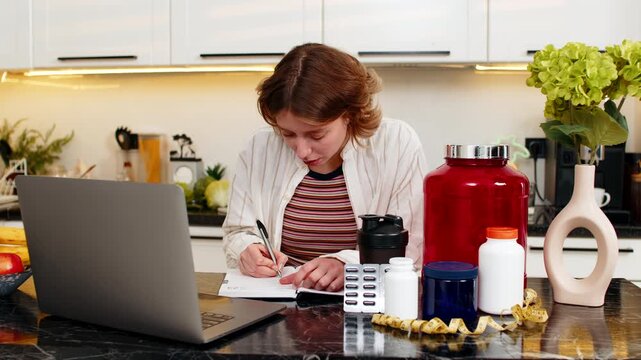 Young woman smiling while writing fitness supplement plan in notebook for diet control and workout goals. Adult girl makes careful nutrition choices for training routine during healthy home planning.