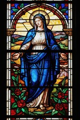 Fototapeta premium Mary stained glass window shows blue robed figure with open hands in garden. Religious art depicts holy mother with roses below landscape scene. Jesus mother symbol of peace and hope.