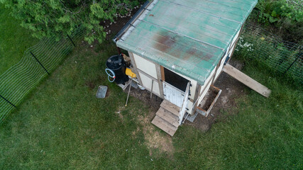 A high-angle drone photograph looking down at a rustic, wooden backyard chicken coop with an open door, wooden ramp, a rainwater collection barrel, and a wire fence enclosure on a green lawn. © Matt