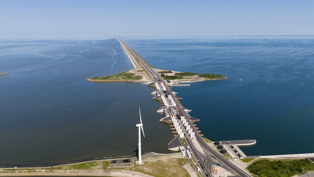 Aerial view of the Afsluitdijk dam featuring a wind turbine and sluice gates along the highway under a clear blue sky in Breezanddijk, Friesland, Netherlands.
