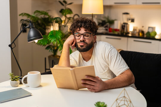 Arabian man at home reads dull book eyes heavy leans on hand slow page turn bored. Egyptian guy sitting at table keeps reading to finish chapter tries focus blinks often breath calm quiet night still