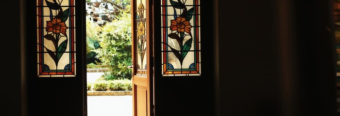 Fototapeta premium Open wooden doors reveal stained glass panels with floral art. Sunlight streams through into a rich green garden. This view offers a peaceful transition between interior and exterior nature.