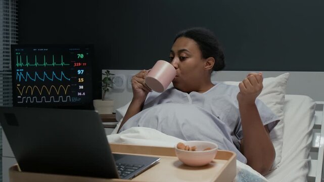 Pregnant Black woman lying in hospital bed,eating cookies and drinking from mug while watching laptop. Footage is suitable for maternity care and prenatal health promos