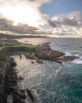 Aerial view of O Vixiador featuring rugged rocky cliffs, crashing ocean waves, and a coastal path under a dramatic cloudy sky, Arteixo, A Coruna, Spain.