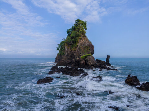 Aerial view of a rocky sea stack covered in lush green trees surrounded by dark volcanic rocks and crashing white waves in the blue ocean under a clear sky.