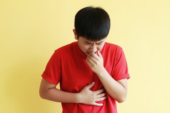 Young boy feels sick and holds his mouth and stomach. He looks unwell and uncomfortable, possibly with nausea or stomach pain. Isolated on yellow background.