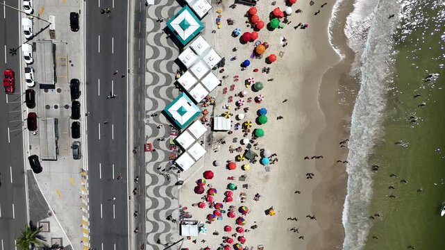 Top down aerial drone footage tracking along Copacabana Beach, following a vehicle beside the iconic wave-pattern promenade, colorful beach umbrellas, sandy shoreline and ocean waters in Rio de Janeir