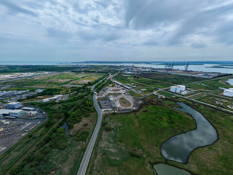 Aerial view of the Grain LNG terminal with industrial storage tanks, green wetlands, and the Thames Estuary under a cloudy sky in Wallend, England, United Kingdom.