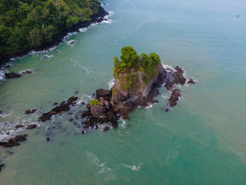 Aerial view of Pantai Karang Agung featuring a rocky sea stack topped with lush green trees surrounded by turquoise water and dark coastal rocks in Ayah, Central Java, Indonesia.