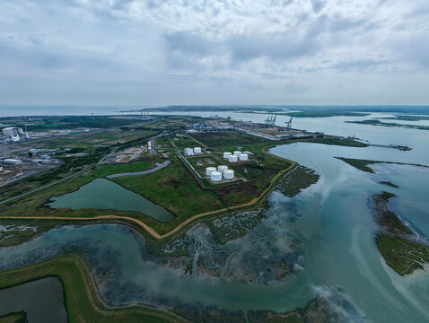 Aerial view of Grain LNG terminal featuring white storage tanks surrounded by green fields and tidal wetlands under a cloudy sky in Wallend, England, United Kingdom.