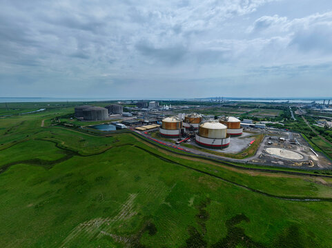 Aerial view of Grain LNG terminal with large storage tanks surrounded by green fields and coastal landscape under a cloudy sky Wallend, England, United Kingdom.