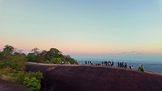 A breathtaking sunrise panorama captures tourists gathered on the massive whale shaped cliffs of Three Whale Rock, overlooking misty mountains and endless tropical wilderness.