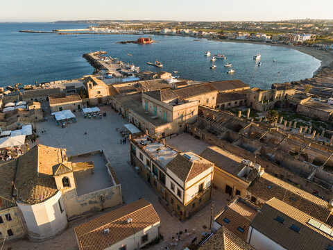Aerial view of the historic Tonnara di Marzamemi and Piazza Regina Margherita with stone buildings and the sea at sunset in Marzamemi, Sicily, Italy.