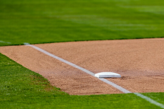 A close-up view of third base on a well-maintained baseball field shows a crisp, white base anchored firmly in a smooth expanse of freshly groomed red clay. 