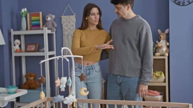 Couple stands by baby crib and hanging mobile as man extends his hand palm out in a nursery inside a building; anticipation parenthood.