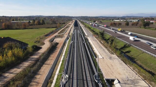 Aerial view of new railway tracks running parallel to a busy highway with trucks and traffic through the green countryside in Desenzano del Garda, Lombardy, Italy.
