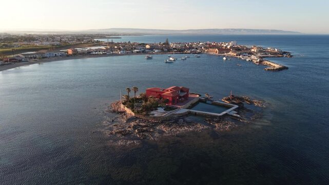 Aerial view of the red villa on Isolotto Brancati surrounded by the calm blue Mediterranean Sea near the historic fishing village of Marzamemi, Sicilia, Italy.