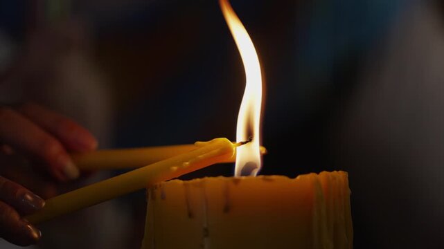 Close up of a burning candle flame against a dark background as blurred worshippers smoothly alternate to pass the fire in calm mindfulness.