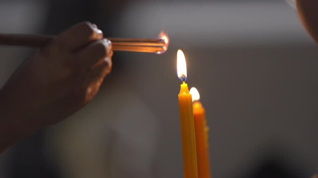 Close up of hands carefully holding a bundle of incense sticks to catch the flame from a small burning yellow candle.