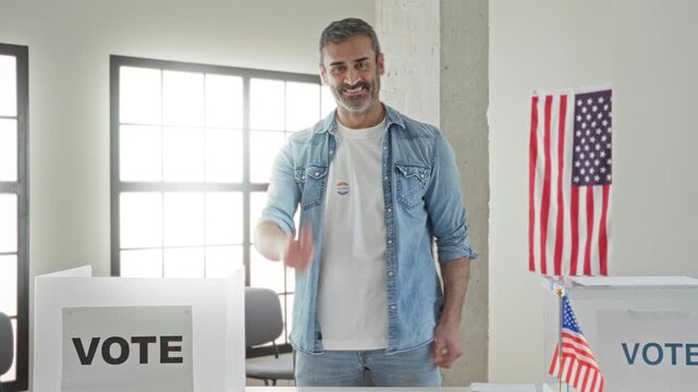 Man giving thumbs up beside vote booth and clear ballot box wearing denim shirt and i voted sticker in a building; civic pride participation.