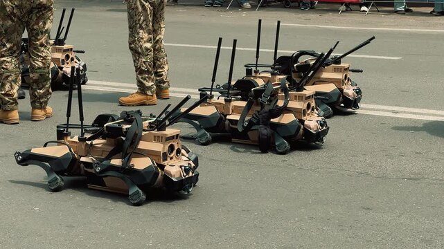 Four quadruped military robotic dogs sit lined up on a city street while police officers and soldiers walk past during an outdoor public technology and defense exhibition event.

