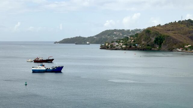 Static wide shot of two small fishing boats anchored in a calm Caribbean bay with lush green tree-covered hills and a small coastal settlement in the background in Puerto Rico. Shot on iPhone 17 Pro.
