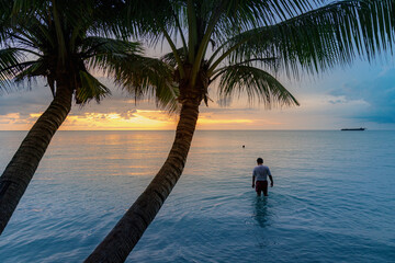 Fototapeta premium A man wades into the ocean at sunset in Koh Kood, Thailand, enjoying the tranquil evening waters. A cargo ship is visible on the horizon.