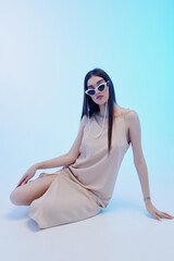 Naklejka premium Young woman in a beige dress and white sunglasses poses seated on a light blue background. Concept of beauty, fashion, and modern style with elegant minimalism.