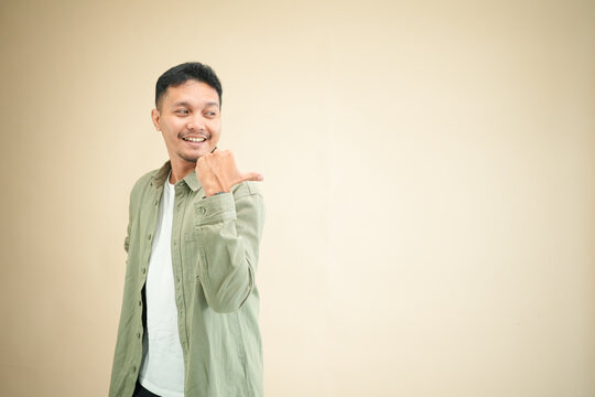 Smiling young Asian man pointing sideways with thumb and laughing, directing attention to copy space in casual olive green shirt and beige studio