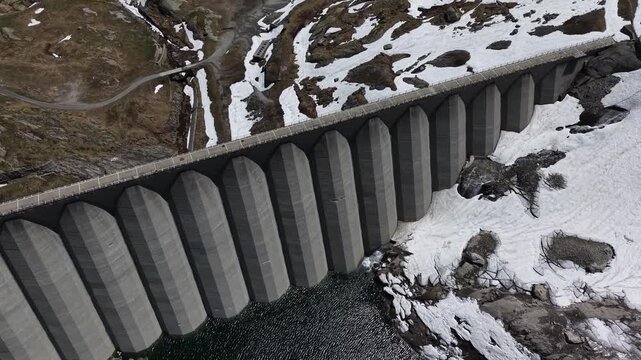 Aerial view of Lucendro Dam featuring concrete arched buttresses, dark reservoir water, and patches of melting snow on the rocky landscape in Airolo, Ticino, Switzerland.