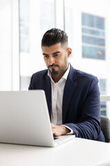 Fototapeta premium Concentrated Indian businessman in formal suit working on laptop in modern office, responding on client e-mails, reviewing business documents, analyzing data, writing reports, preparing presentation