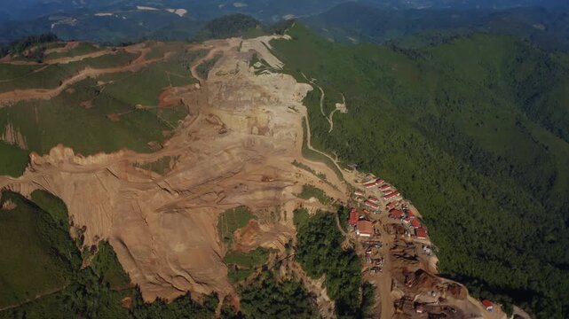 Top-down aerial tracking shot of an active iron ore mine, Red laterite soil and mining equipment contrast with surrounding green forest and mountains in Xieng Khouang province, Lao PDR.