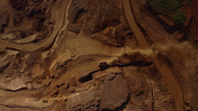 Top-down aerial tracking shot of an active iron ore mine, Red laterite soil and mining equipment contrast with surrounding green forest and mountains in Xieng Khouang province, Lao PDR.