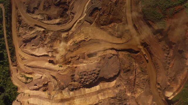 Top-down aerial tracking shot of an active iron ore mine, Red laterite soil and mining equipment contrast with surrounding green forest and mountains in Xieng Khouang province, Lao PDR.