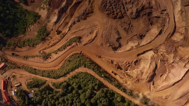 Top-down aerial tracking shot of an active iron ore mine, Red laterite soil and mining equipment contrast with surrounding green forest and mountains in Xieng Khouang province, Lao PDR.