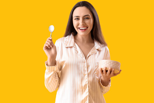 Young woman in pajamas with spoon and bowl on yellow background