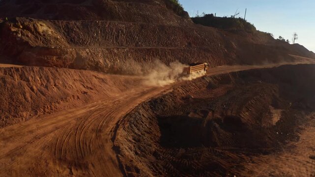 Top-down aerial tracking shot of an active iron ore mine, Red laterite soil and mining equipment contrast with surrounding green forest and mountains in Xieng Khouang province, Lao PDR.