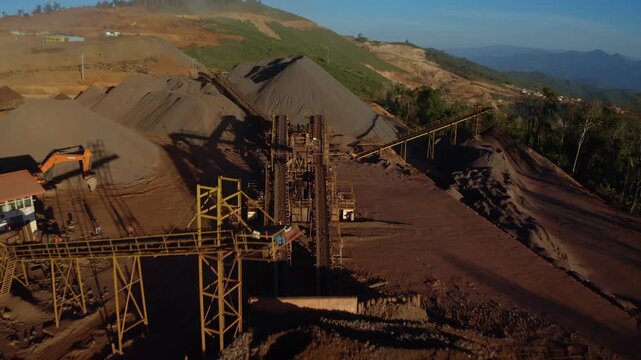 Top-down aerial tracking shot of an active iron ore mine in Xieng Khouang province, Lao PDR. Red laterite soil and mining equipment contrast with surrounding green forest and mountains.