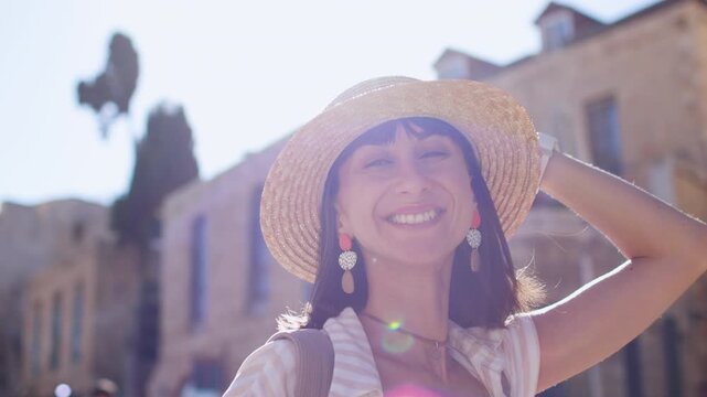 A young woman in a straw hat and summer dress holds the camera in front of her face, looking up at a sunlit historic town square. She stands in the center of the frame
