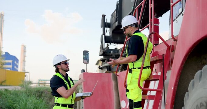 Adult Caucasian male logistics worker passes laptop to colleague on container truck ladder, confirming shipment record, safety checklist, freight loading status, yard coordination during dispatch