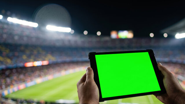 First-person perspective holding a blank green screen tablet, blurred night stadium background. Tailored for live sports analytics interfaces, broadcasting apps, and event management mockups.