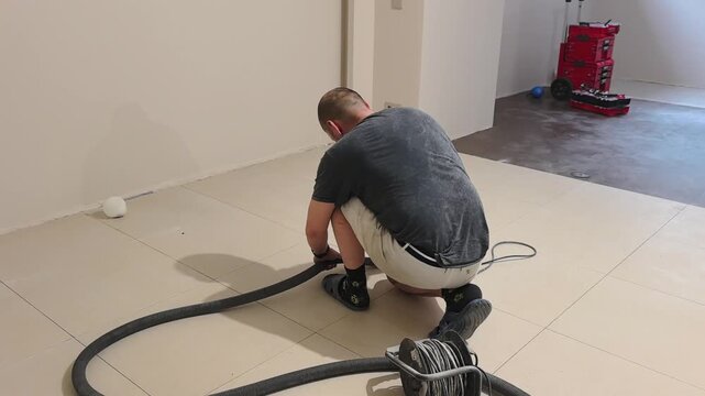 A man cleans the grout in porcelain tiles after laying the tiles, using an angle grinder and a vacuum cleaner to vacuum the floor in an empty room. The work is being done during the day at a construct