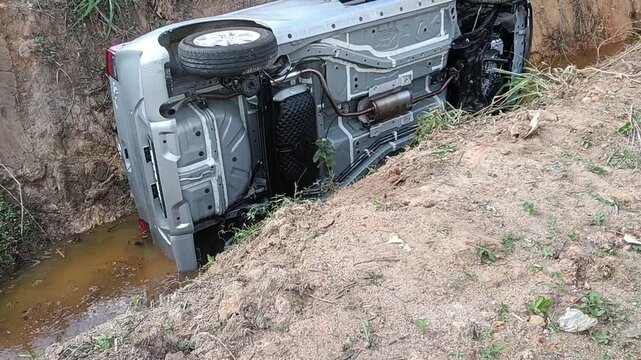 A silver car lies on its side in a muddy ditch beside a dirt road, with the hood open. It appears to be a rural accident scene in malaysia with no visible responders.