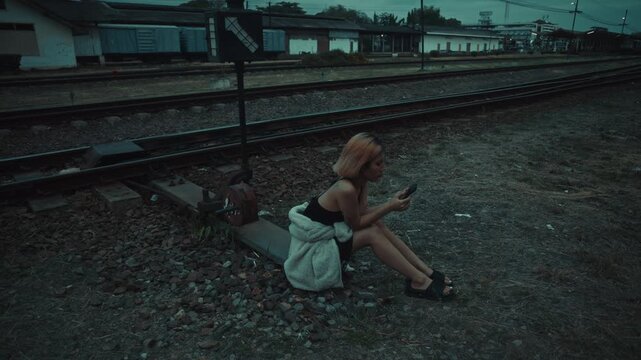 Young woman sitting on gravel beside railway tracks at dusk