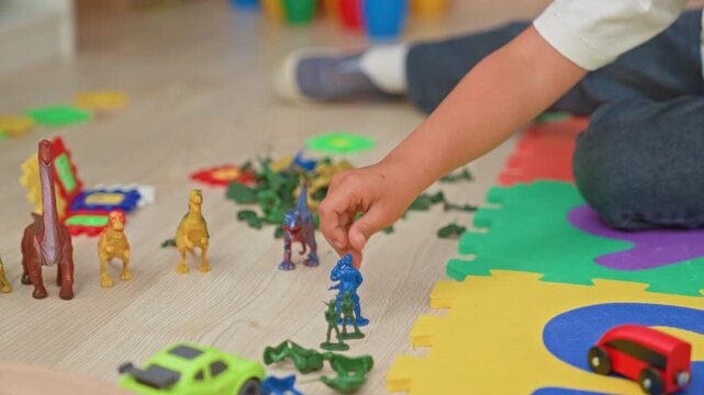 Boy sitting on a foam puzzle mat arranging toy dinosaurs, plastic soldiers and cars, hand picking up a blue soldier on wooden floor inside a building; playfulness imagination.