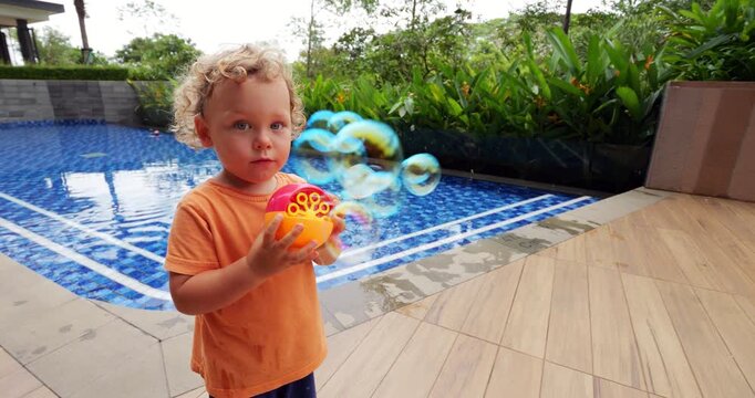 Young child explores his new toy - small soap bubble generator, captured in mesmerizing slow motion. With wide-eyed fascination, he holds tiny machine as colorful bubbles rush out, soaring towards