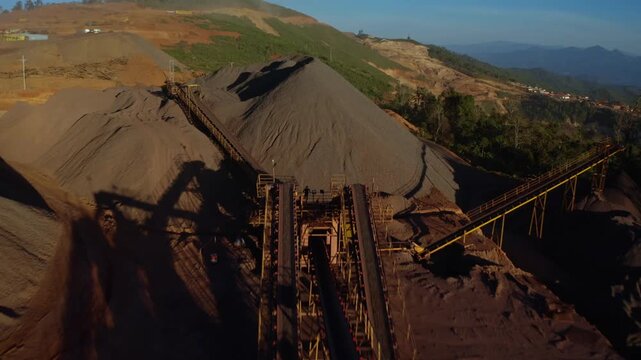 Top-down aerial tracking shot of an active iron ore mine, Red laterite soil and mining equipment contrast with surrounding green forest and mountains in Xieng Khouang province, Lao PDR.